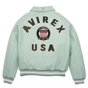 Może przedstawiać: Jasnozielona sk&oacute;rzana kurtka typu varsity z czarnym napisem AVIREX wygiętym w łuk nad okrągłą naszywką z amerykańską flagą i napisem ALL AMERICANS ESTABLISHED 1975, z czarnym napisem USA umieszczonym pod naszywką oraz prążkowanymi ściągaczami.