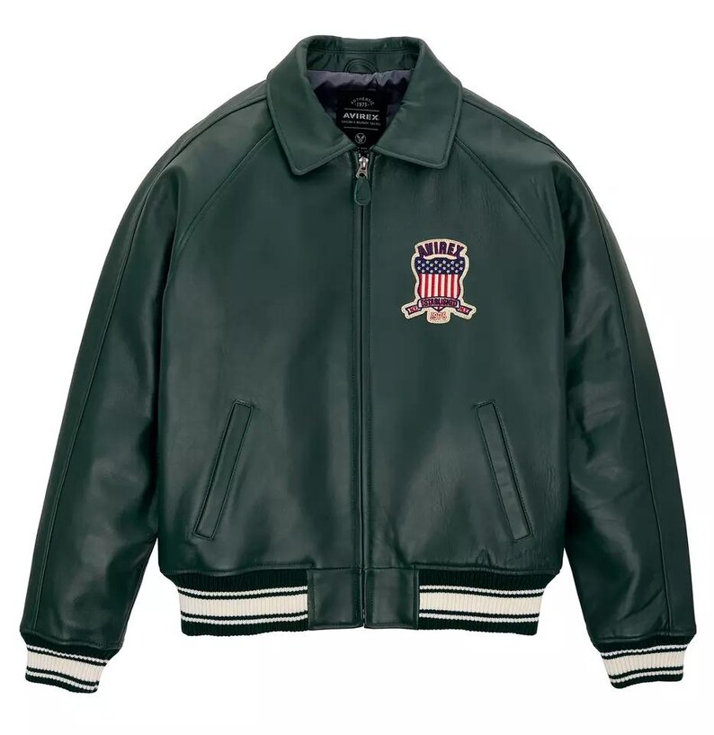 Puede incluir: Una chaqueta de cuero verde oscuro con cremallera frontal completa, cuello cl&aacute;sico y pu&ntilde;os y dobladillo acanalados con rayas blancas. Un parche en el pecho izquierdo muestra el texto AVIREX con un gr&aacute;fico de escudo que contiene estrellas.