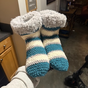 May include: Hand-crocheted slipper socks in teal, cream, and gray stripes with a plush gray cuff. These cozy socks are designed for indoor wear and provide warmth and comfort. The handmade design adds a unique touch to the product.
