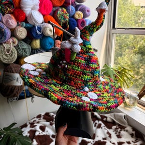 May include: A crocheted hat featuring a vibrant, multi-colored yarn base with a pointed, curved top. The hat is decorated with several small, white, three-dimensional crocheted mushroom shapes attached to the crown and the wide, flat brim.