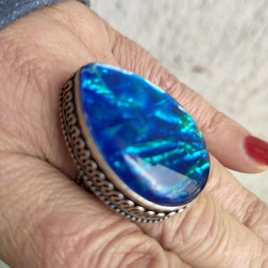 Puede incluir: Un llamativo anillo con una gran ópalo azul ovalado, que muestra destellos iridiscentes verdes y azules. El ópalo está engastado en una banda de metal plateado con un borde detallado y texturizado. Una pieza de joyería audaz, el anillo es un accesorio llamativo.