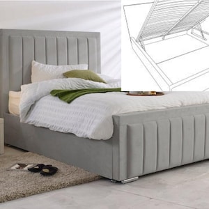 May include: A grey upholstered bed with a tall, vertically-panelled headboard. The bed has a storage base and is covered with white bedding and a green throw. A small side table with a lamp is visible.