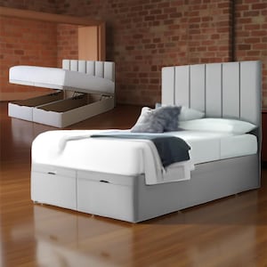 May include: A light grey upholstered bed with a tall, vertically-panelled headboard. The bed has a storage base with two drawers and a mattress. A second bed is shown with the mattress raised, revealing storage compartments. Pillows and a blanket are on the bed.