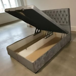 May include: A grey upholstered storage bed with the lid open, showing the storage area. The headboard features a diamond pattern. The rectangular bed frame is on a light-coloured floor.