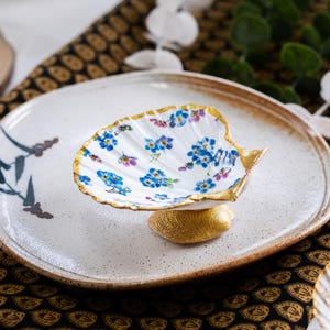 May include: A decorative ceramic seashell-shaped dish with a gold-painted rim and base, filled with blue and purple floral designs. The dish sits on a speckled white plate with a brown rim, accented with painted floral details.