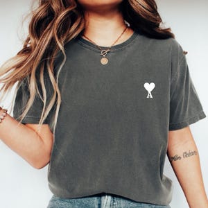 Puede incluir: Camiseta gris oscuro con un corazón blanco y la letra "A" estampada en el pecho. La camiseta tiene cuello redondo y mangas cortas. La modelo lleva un collar y una pulsera dorados.