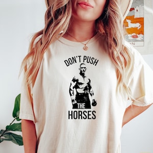 Puede incluir: Camiseta color crema con un gráfico negro de una figura musculosa y el texto "DON'T PUSH THE HORSES" en negrita. El diseño está centrado en la parte delantera de la camiseta.