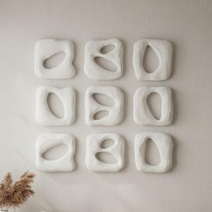 May include: A set of nine square white wall art pieces arranged in a three-by-three grid. Each piece features a smooth, rounded frame with an irregular, organic cutout in the centre, creating a cohesive collection of textured, sculptural wall decor.