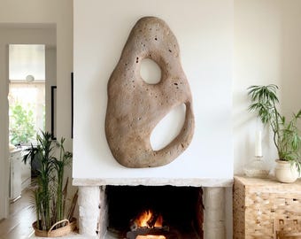 Pode incluir: Uma grande escultura de parede abstrata em pedra, com uma forma natural e orgânica. A escultura é de cor castanho claro e tem uma grande abertura circular e uma abertura curva. Está montada numa parede branca acima de uma lareira.