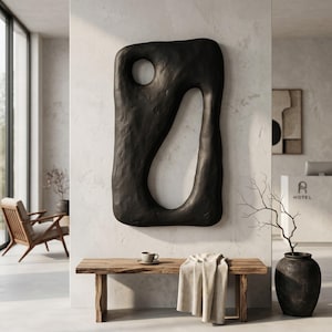 May include: A large, black, rectangular wall sculpture with an irregular, textured surface and two distinct cutouts: a small circular opening at the top and a larger, elongated, organic-shaped void extending through the center of the piece.