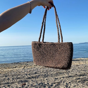 Può includere: Una borsa tote rettangolare marrone all'uncinetto con due manici intrecciati. La borsa è tenuta contro uno sfondo di spiaggia sabbiosa, oceano e cielo blu. La trama della borsa è visibile, mostrando le strette maglie all'uncinetto.