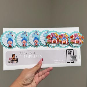 May include: A set of cake toppers featuring a clown and circus theme. The toppers include the number "1" and circus tent designs, with a light blue and white color scheme. The text "PRISCILLA IMPRINTS" is also visible.