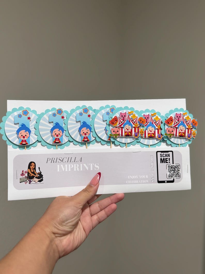 May include: A set of cake toppers featuring a clown and circus theme. The toppers include the number "1" and circus tent designs, with a light blue and white color scheme. The text "PRISCILLA IMPRINTS" is also visible.