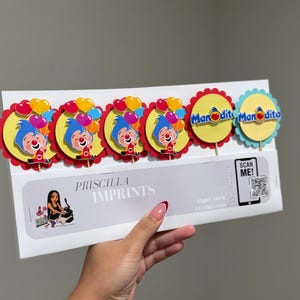 May include: A package of cupcake toppers featuring a cartoon clown with blue hair and a red nose, holding colorful balloons. The toppers are on yellow and red scalloped circles. The package includes the text "Priscilla Imprints".