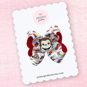 May include: A hair bow made of white grosgrain ribbon featuring a repeating pattern of Curious George and colorful balloons, with a red underside and a central embroidered patch of the character's face on a white display card.