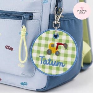 May include: A round, green and white gingham fabric tag attached to a light blue backpack with a metal lobster clasp. The tag features an embroidered yellow and red construction excavator and the name Tatum in blue cursive script embroidery.