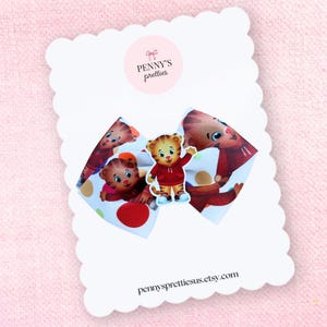 May include: A white and red polka dot hair bow featuring a cartoon tiger character. The bow is attached to a white card with a scalloped edge. The card has the text "penny's pretties" and "pennysprettiesus.etsy.com".