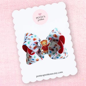 May include: A large white hair bow with a red trim and a cartoon character design. The bow features a central character embellishment and is mounted on a scalloped card with the seller's logo and website.