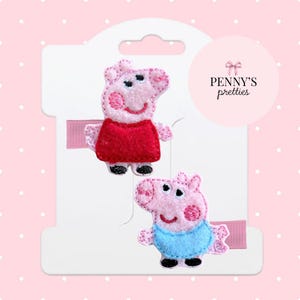 May include: Two pink pig hair clips with embroidered details, one in a red dress and the other in a blue outfit. The clips are attached to a white card with a pink ribbon and the text "Penny's Pretties".