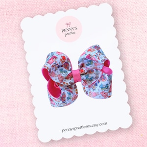 May include: A large, decorative hair bow with a pink center and edges. The bow features a white background with a colorful pattern of Easter-themed illustrations. The bow is attached to a white card with the text "Penny's Pretties" and "pennysprettiesus.etsy.com".