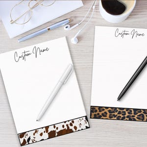 Peut inclure: Deux blocs-notes blancs avec le texte "Custom Name" en écriture noire. L'un des blocs-notes a un motif de peau de vache marron et blanc en bas, et l'autre a un imprimé léopard. Un stylo blanc et un stylo noir sont sur les blocs-notes.