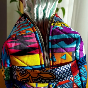 Peut inclure: Un sac à couches matelassé coloré avec un motif patchwork vibrant. Le sac est doté d'une fermeture éclair et d'une sangle à motifs. Des couches blanches sont visibles à l'intérieur du sac ouvert. Le design du sac comprend un mélange de motifs géométriques et abstraits.