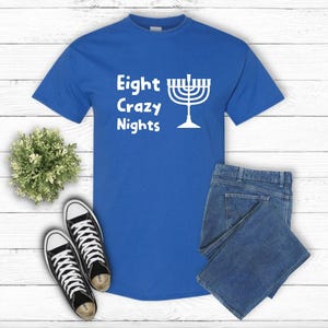 May include: A royal blue t-shirt with the text "Eight Crazy Nights" and a white menorah graphic. The shirt is paired with blue jeans and black and white sneakers, all arranged on a white wooden surface.