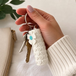 Peut inclure: Accessoire de porte-clés blanc avec un motif de cœurs et un accent floral. L'accessoire est attaché à un porte-clés argenté avec plusieurs clés. L'accessoire est tenu dans une main.