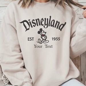 Op de afbeelding: Beige sweatshirt met de zwarte tekst "Disneyland", de afbeelding van Mickey Mouse en de tekst "EST 1955 Your Text". De trui heeft een ronde hals en lange mouwen.