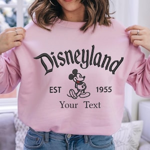 May include: Pink sweatshirt with a black embroidered Disneyland logo, Mickey Mouse, and the text "EST 1955 Your Text".
