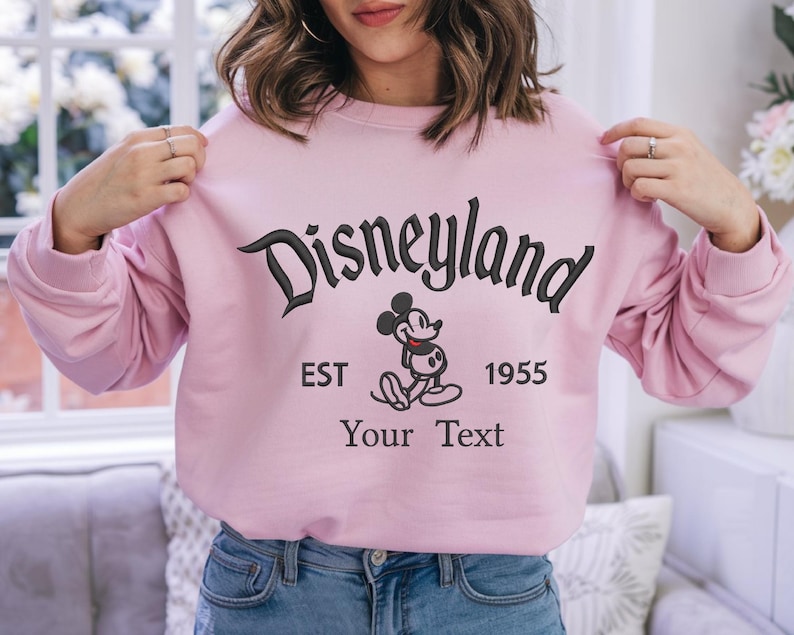 May include: Pink sweatshirt with a black embroidered Disneyland logo, Mickey Mouse, and the text "EST 1955 Your Text".