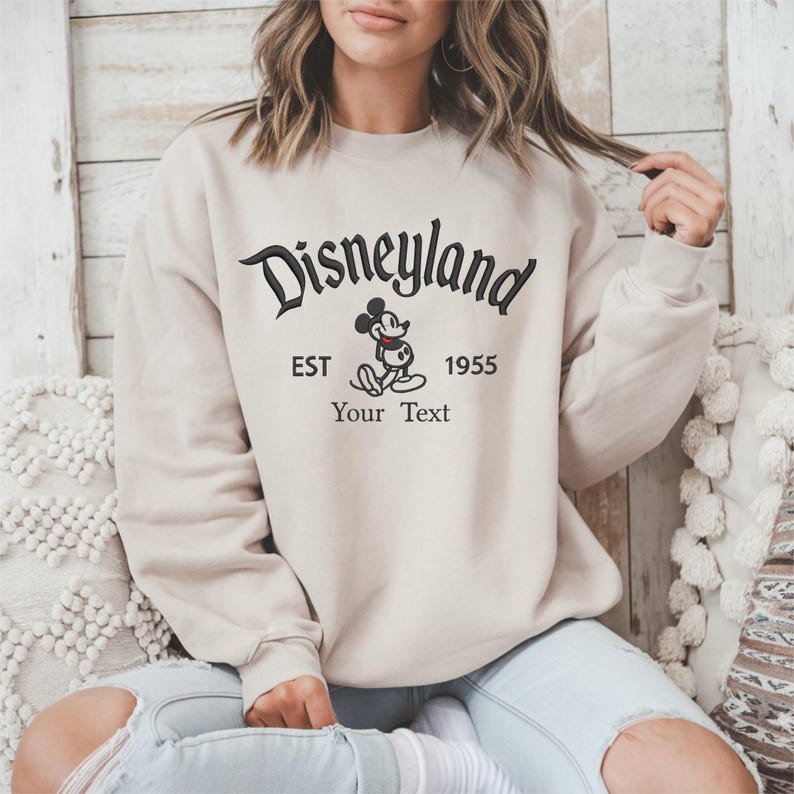Op de afbeelding: Beige sweatshirt met de zwarte tekst "Disneyland", de afbeelding van Mickey Mouse en de tekst "EST 1955 Your Text". De trui heeft een ronde hals en lange mouwen.