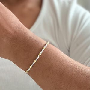 May include: A delicate bracelet with alternating gold and silver beads. The bracelet is worn on a person's wrist, showcasing its simple yet elegant design. The gold beads add a touch of warmth to the piece.