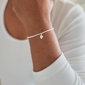 May include: A delicate silver beaded bracelet featuring a small, polished silver heart charm. The bracelet is composed of tiny, uniform metallic spheres strung together to form a continuous band, centered with a single dangling heart-shaped pendant.