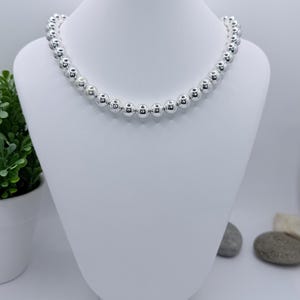 May include: A silver beaded necklace displayed on a white mannequin bust. The necklace features numerous round, shiny beads. The bust is set against a white background, with a small plant and stones in the background.