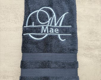 May include: A navy blue hand towel with the embroidered initials "OM" above the name "Mae" in a light blue script. The towel has a textured surface and a decorative band near the bottom.