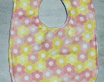May include: A baby bib featuring a repeating hexagonal floral pattern in shades of soft pink, pale yellow, and white. The bib has a rounded shape with a neck opening and a single plastic snap closure at the top, finished with white stitching.