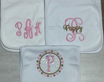 May include: Three white baby burp cloths with embroidered designs. The top left cloth features a pink monogram. The top right cloth displays a pink monogram with the name Poppy in gold thread. The bottom cloth shows a pink letter P in a circle.