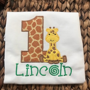 May include: White onesie with a brown and orange giraffe number one and a yellow giraffe with brown spots. The name "Lincoln" is embroidered in green below the giraffe.