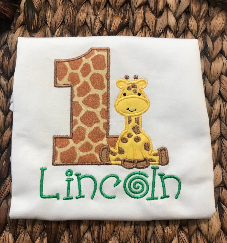 May include: White onesie with a brown and orange giraffe number one and a yellow giraffe with brown spots. The name "Lincoln" is embroidered in green below the giraffe.