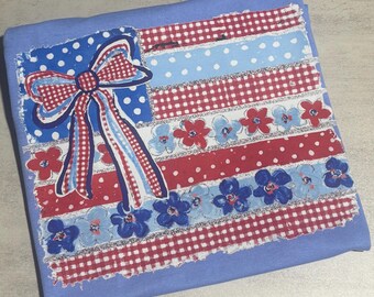 Puede incluir: Un textil azul claro con un diseño patriótico. El diseño presenta un gran lazo y rayas en rojo, blanco y azul, con un estampado floral. El artículo está doblado, lo que sugiere que es un textil decorativo.
