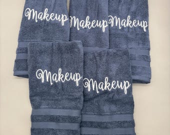 Puede incluir: Cinco toallas rectangulares de tela de rizo azul oscuro dispuestas en grupo. Cada toalla presenta la palabra Makeup escrita en letras cursivas blancas en el centro. Las toallas tienen una superficie de tela texturizada y bandas acanaladas.