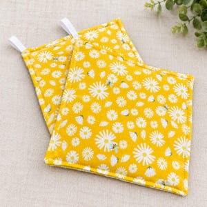 May include: Two square quilted potholders in a vibrant yellow fabric featuring a repeating pattern of white daisy flowers and small bees. Each potholder includes a white fabric loop attached to one corner for convenient hanging storage.