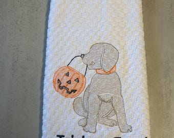 May include: A white waffle-weave kitchen towel featuring an embroidered gray puppy wearing an orange collar. The puppy is holding an orange jack-o'-lantern pumpkin bucket in its mouth. The text "Trick or Treat" is embroidered in black below the dog.