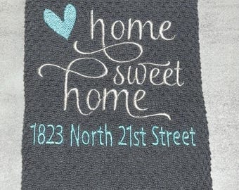 Puede incluir: Un paño de cocina gris oscuro texturizado con texto bordado en blanco en cursiva que dice home sweet home, un icono de corazón azul claro y la dirección 1823 North 21st Street en letras de bloque azul claro en la parte inferior.