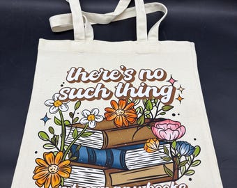 Puede incluir: Una bolsa de tela color crema con una ilustración gráfica de una pila de tres libros rodeada de flores coloridas. El texto "there's no such thing as too many books" está impreso en una fuente retro de color marrón sobre y debajo de los libros.