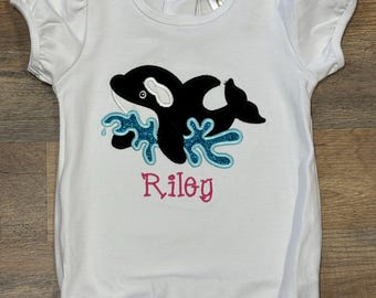 Puede incluir: Romper blanco para bebé con mangas cortas y dobladillo con volantes. El romper presenta una aplicación de orca en blanco y negro con salpicaduras de agua azules y el nombre "Riley" bordado en rosa.