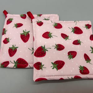 May include: Two pink and white pot holders with a strawberry pattern. The pot holders have white polka dots and red strawberries with green leaves.