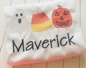 May include: A white t-shirt featuring embroidered Halloween designs including a white ghost, a yellow, orange, and white candy corn, and an orange jack-o'-lantern. The name Maverick is embroidered in dark gray lettering below the designs.