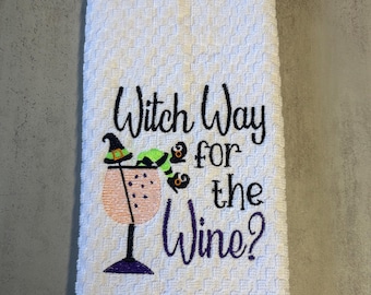 May include: A white waffle-weave kitchen towel featuring black embroidered text that reads Witch Way for the Wine? The design includes a wine glass with a witch hat and green striped legs hanging over the rim, stitched in black, green, and purple.